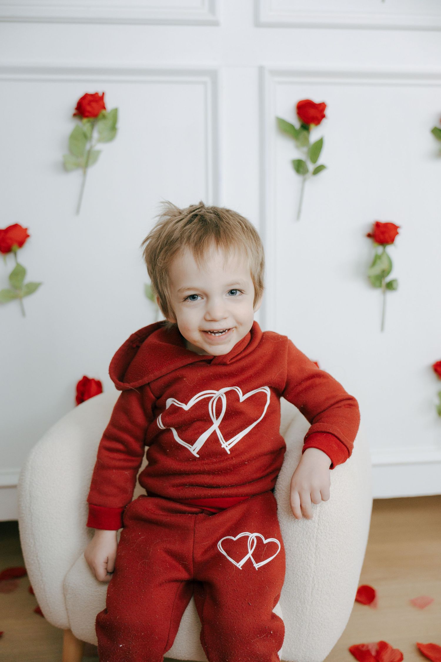 A small child wearing a red Valentine's Day themed outfit sits in a white chair surrounded by red roses.