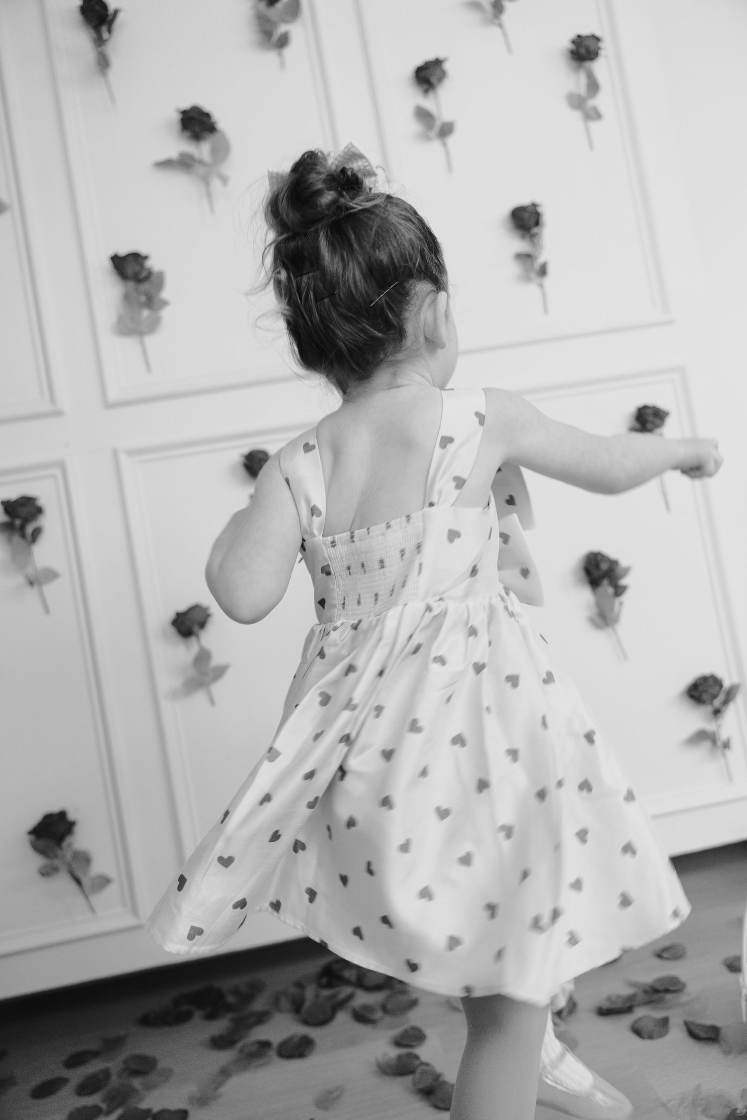 A black and white photo of a twirling child in a patterned dress against a wall decorated with roses.