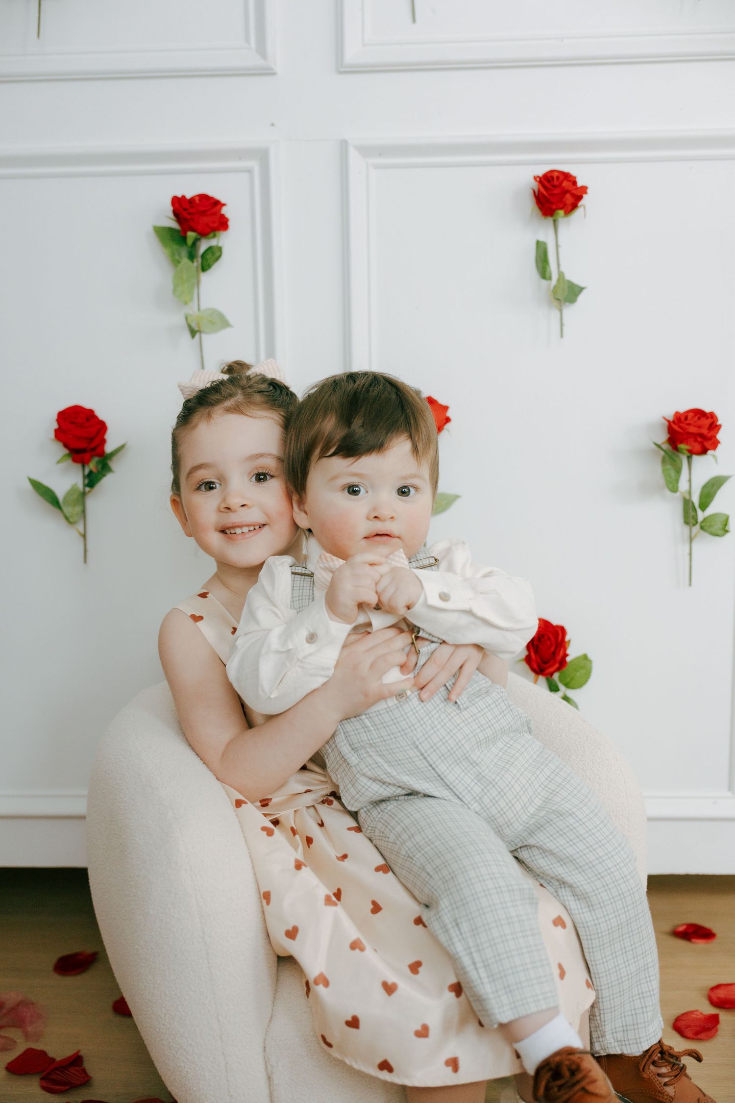 Children sitting together on white chair with red roses decorating white wall behind them.