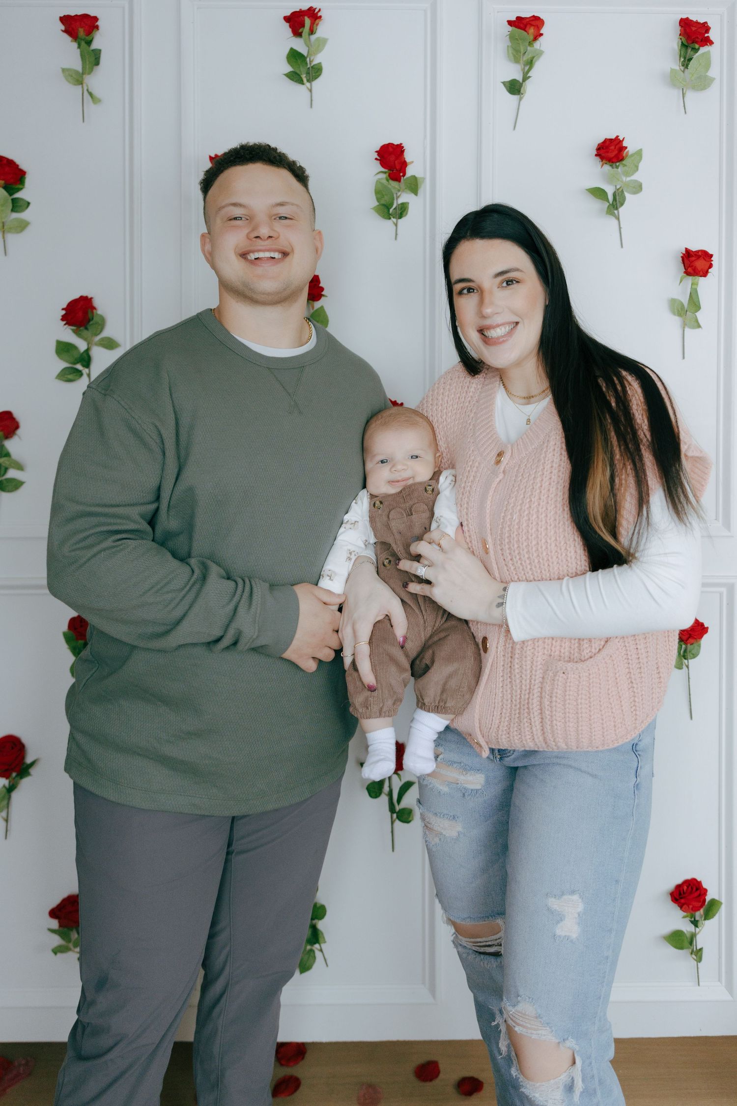Family photo session with red roses backdrop showing couple in casual attire holding baby against white wall.