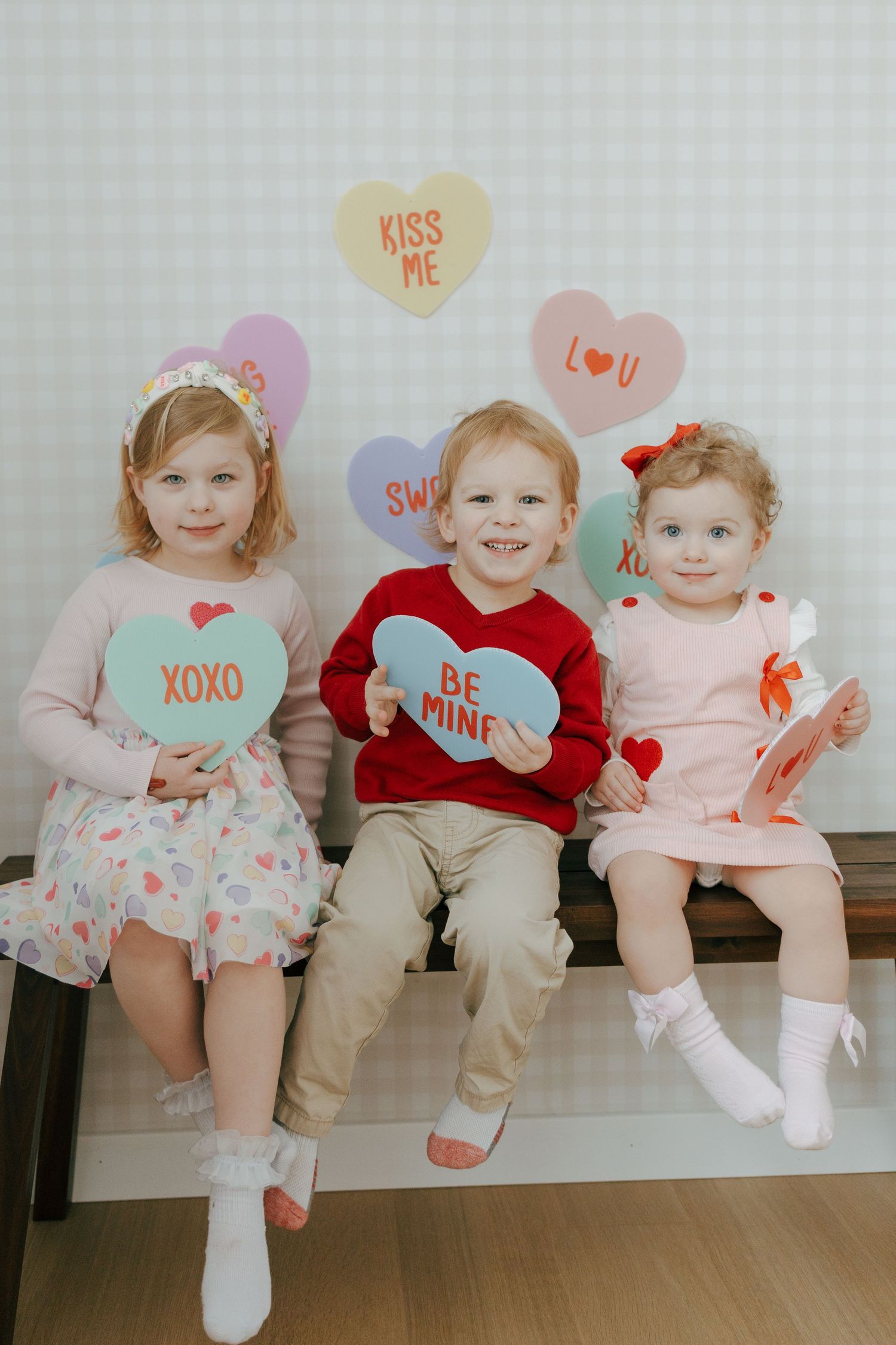 Three toddlers sit on a white bench wearing Valentine's Day themed outfits with heart decorations above their heads.