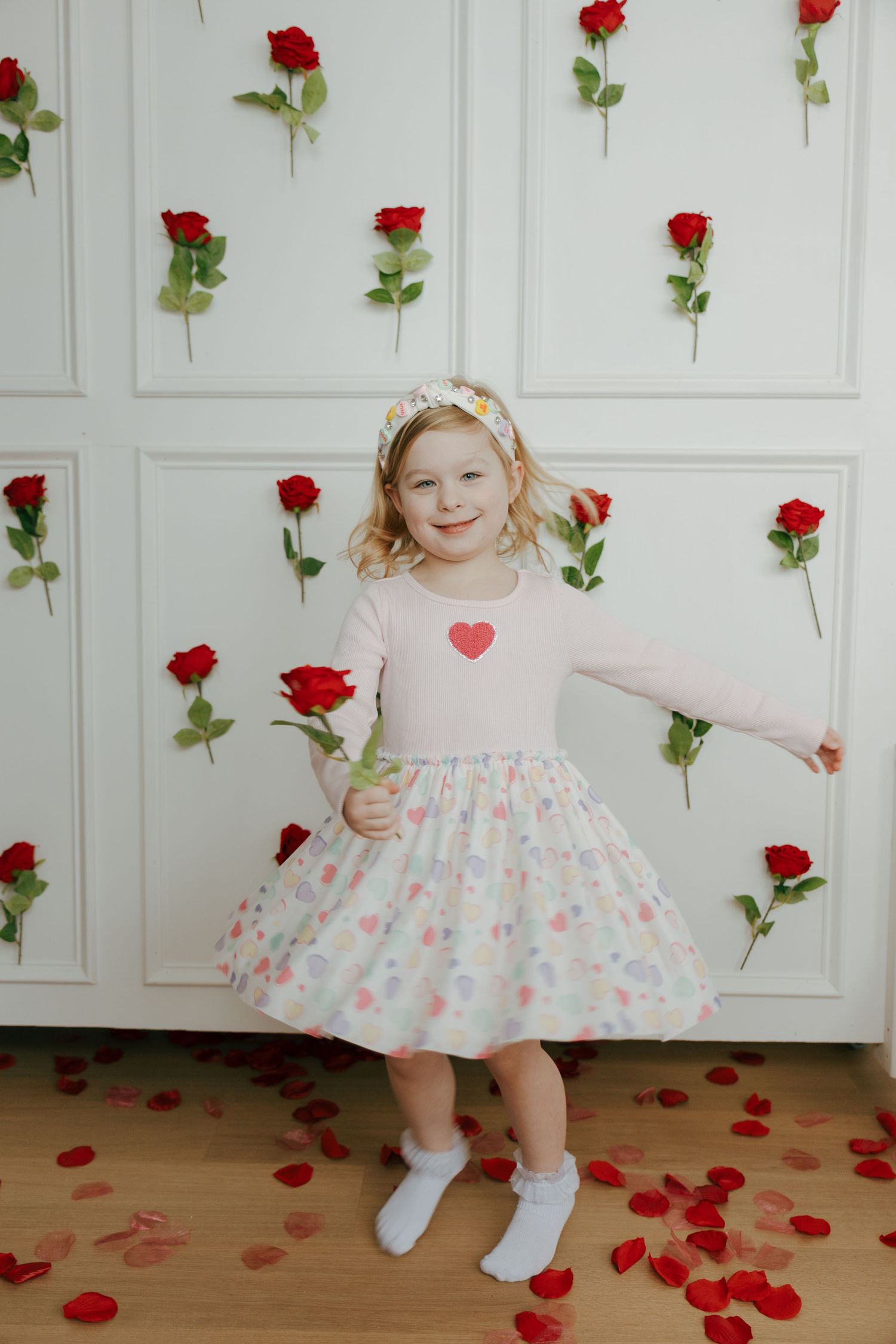 Young child in pink dress dances and twirls among red rose petals and flowers against white paneled wall backdrop.