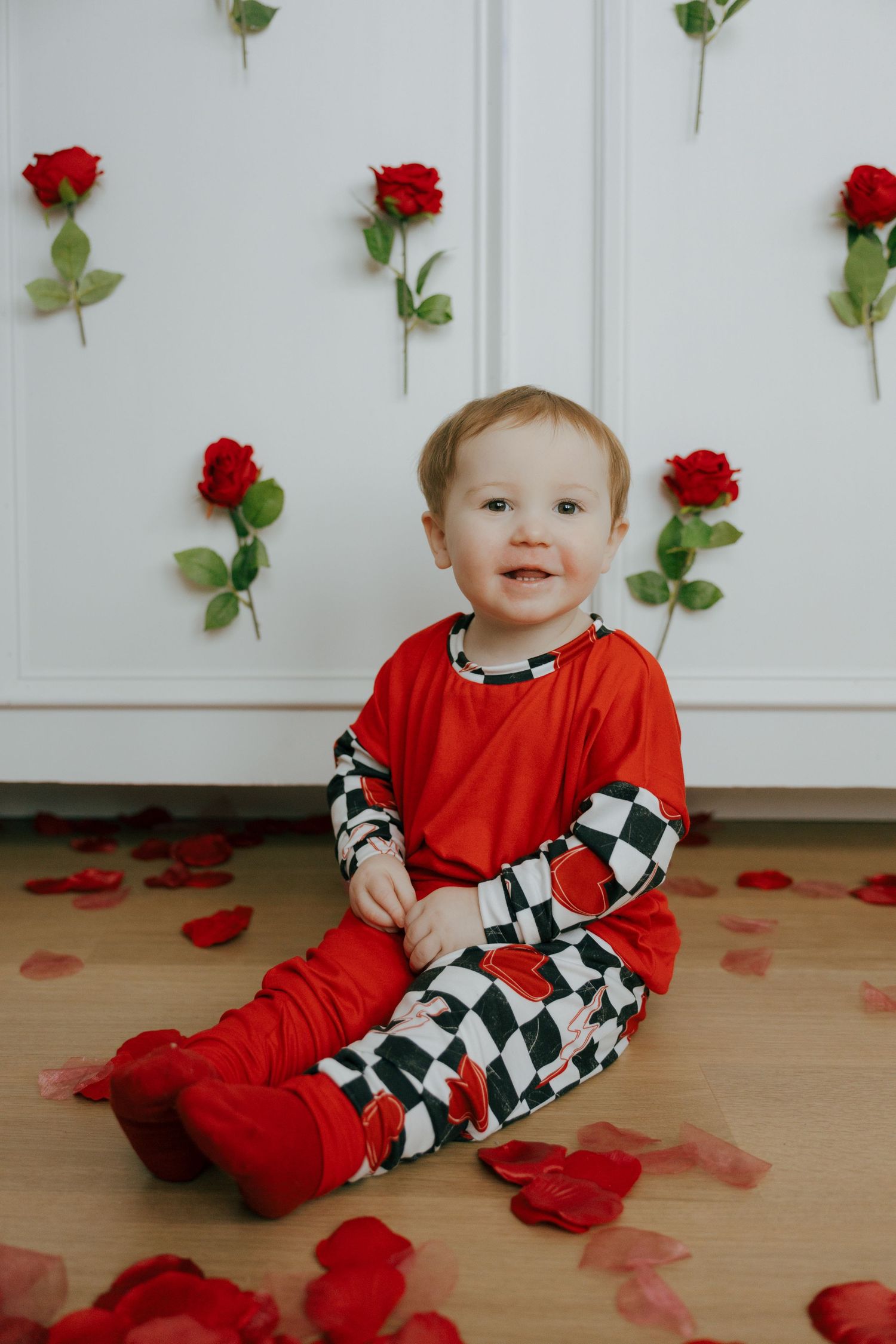 Small child in red outfit sits on wooden floor with scattered red rose petals and roses on wall behind them.