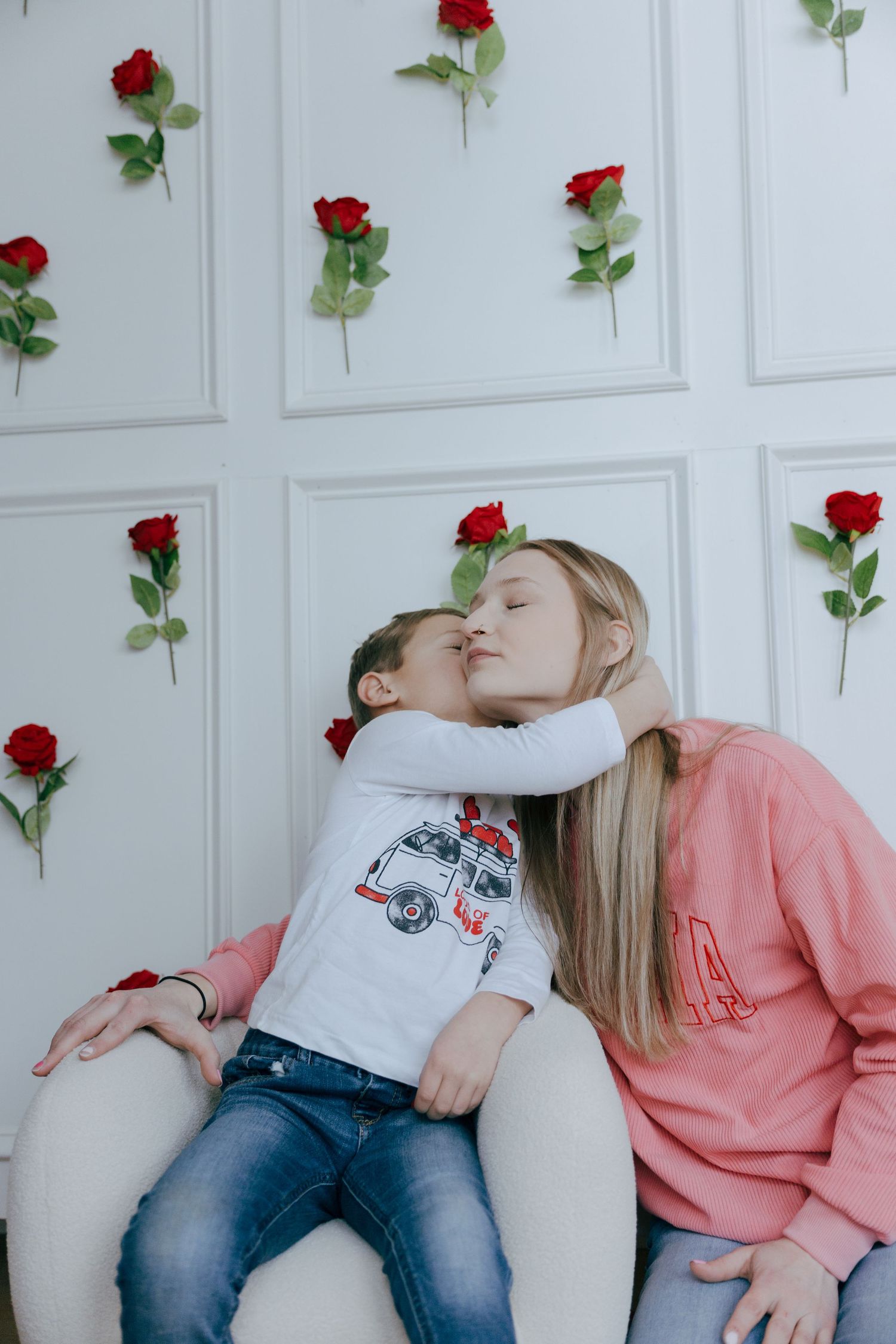 A romantic couple shares tender kisses while sitting against a white wall decorated with red roses.