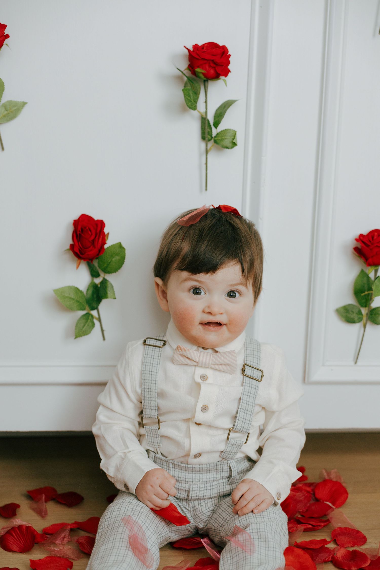 Baby in gray overalls and white shirt sits on floor surrounded by red rose petals against white paneled wall.
