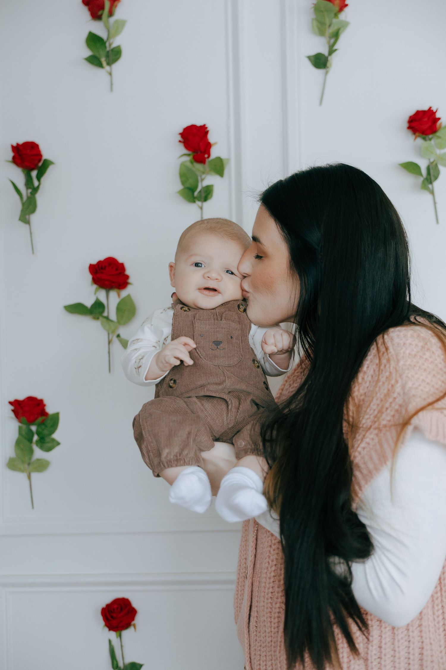 Mother in peach sweater shares tender moments with baby against white wall decorated with red roses.