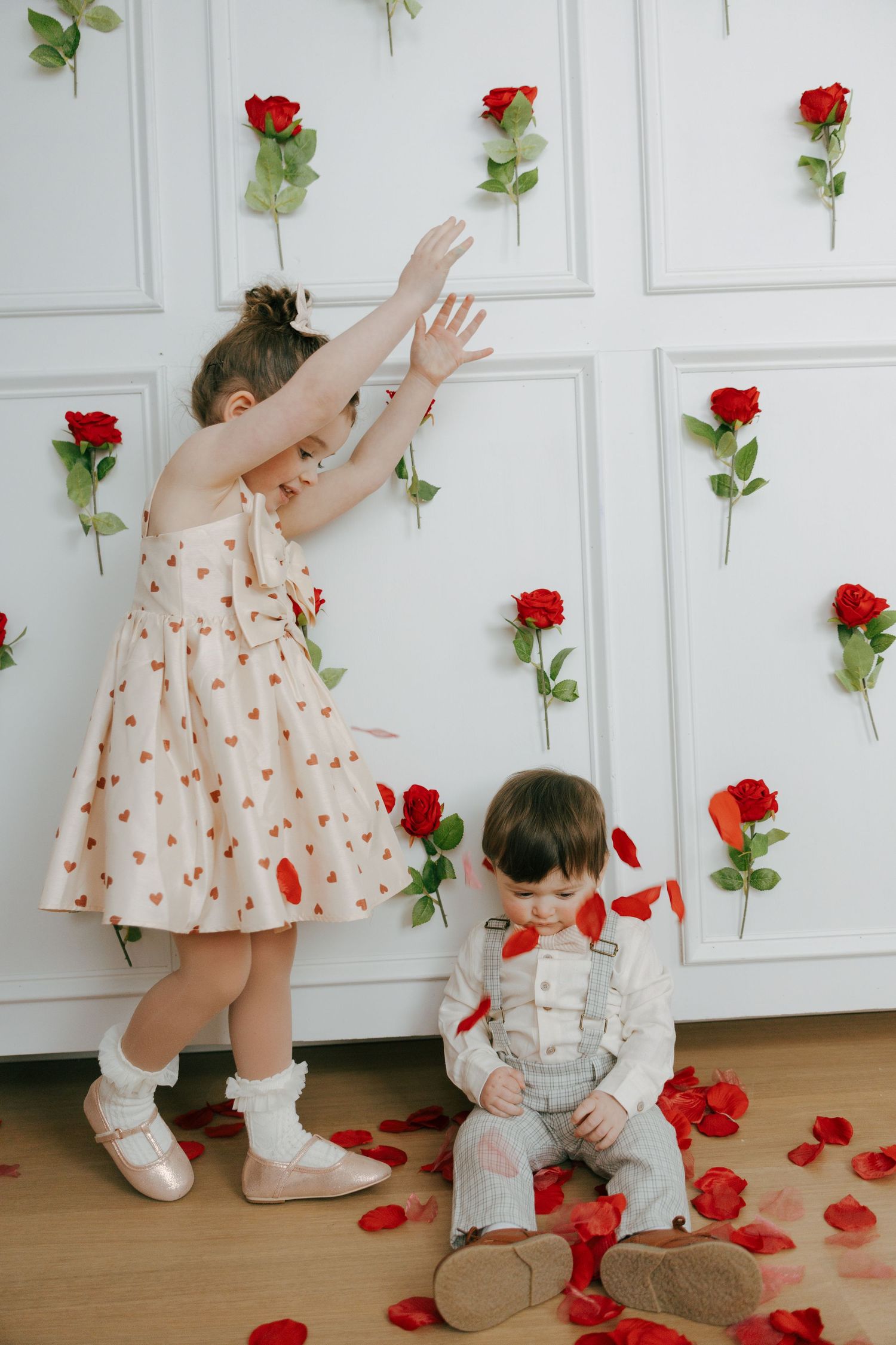 Children in polka dot dresses play with red roses against a white wall in a sweet photo series.
