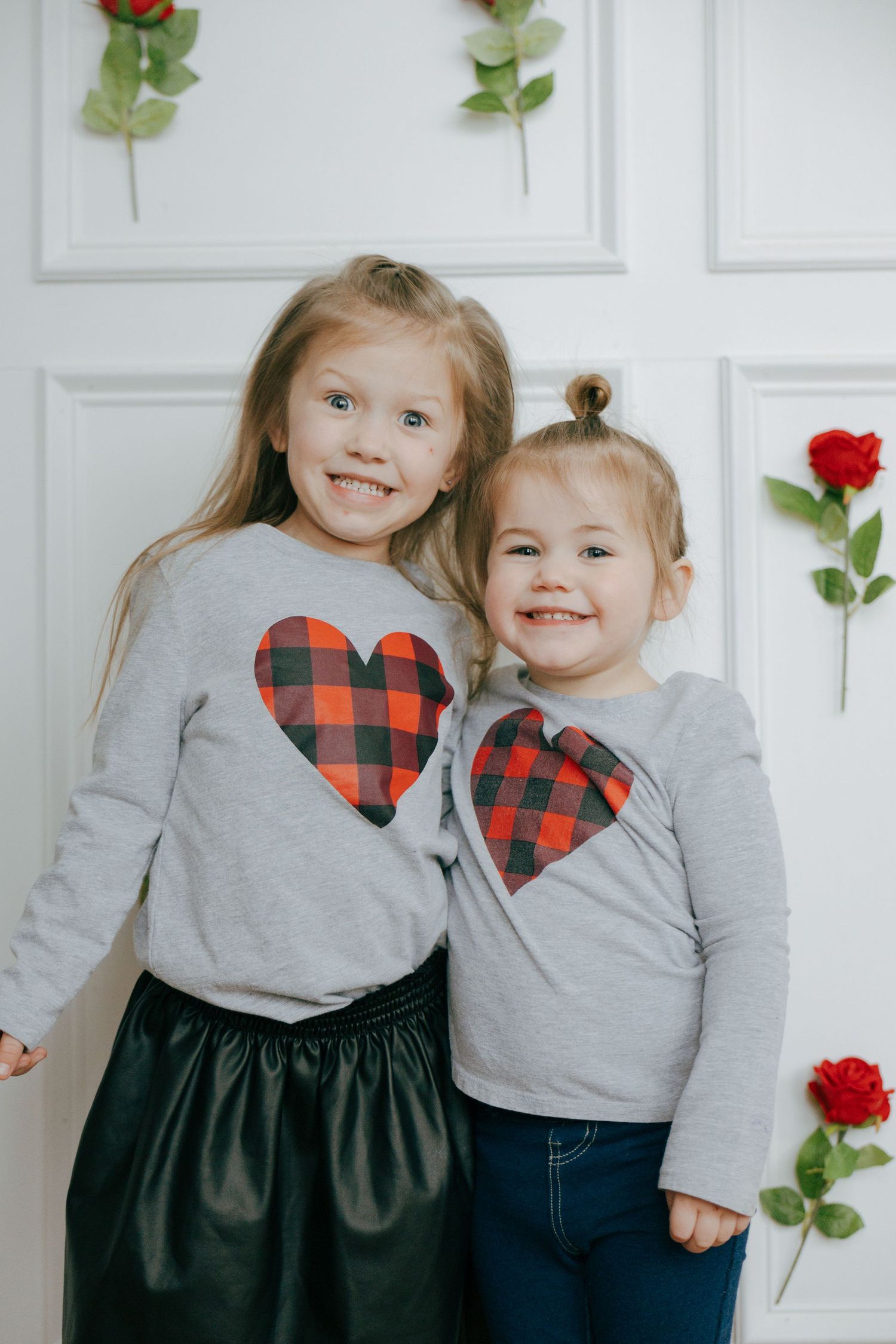 Two children in matching gray sweaters with buffalo plaid hearts and dark bottoms pose together against a white wall.