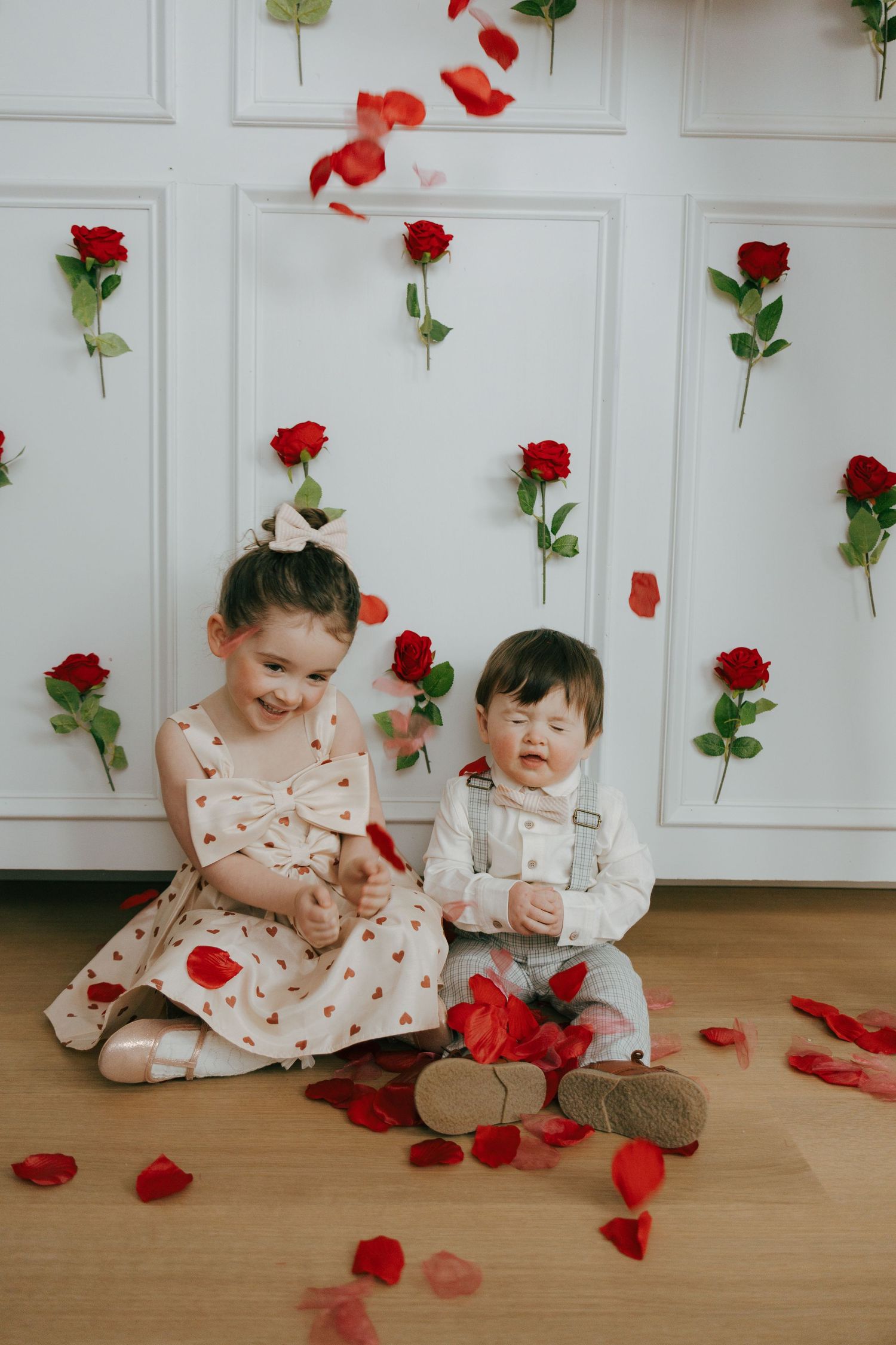 Two children sit on a hardwood floor surrounded by red roses and petals against a white wall with floral decorations.