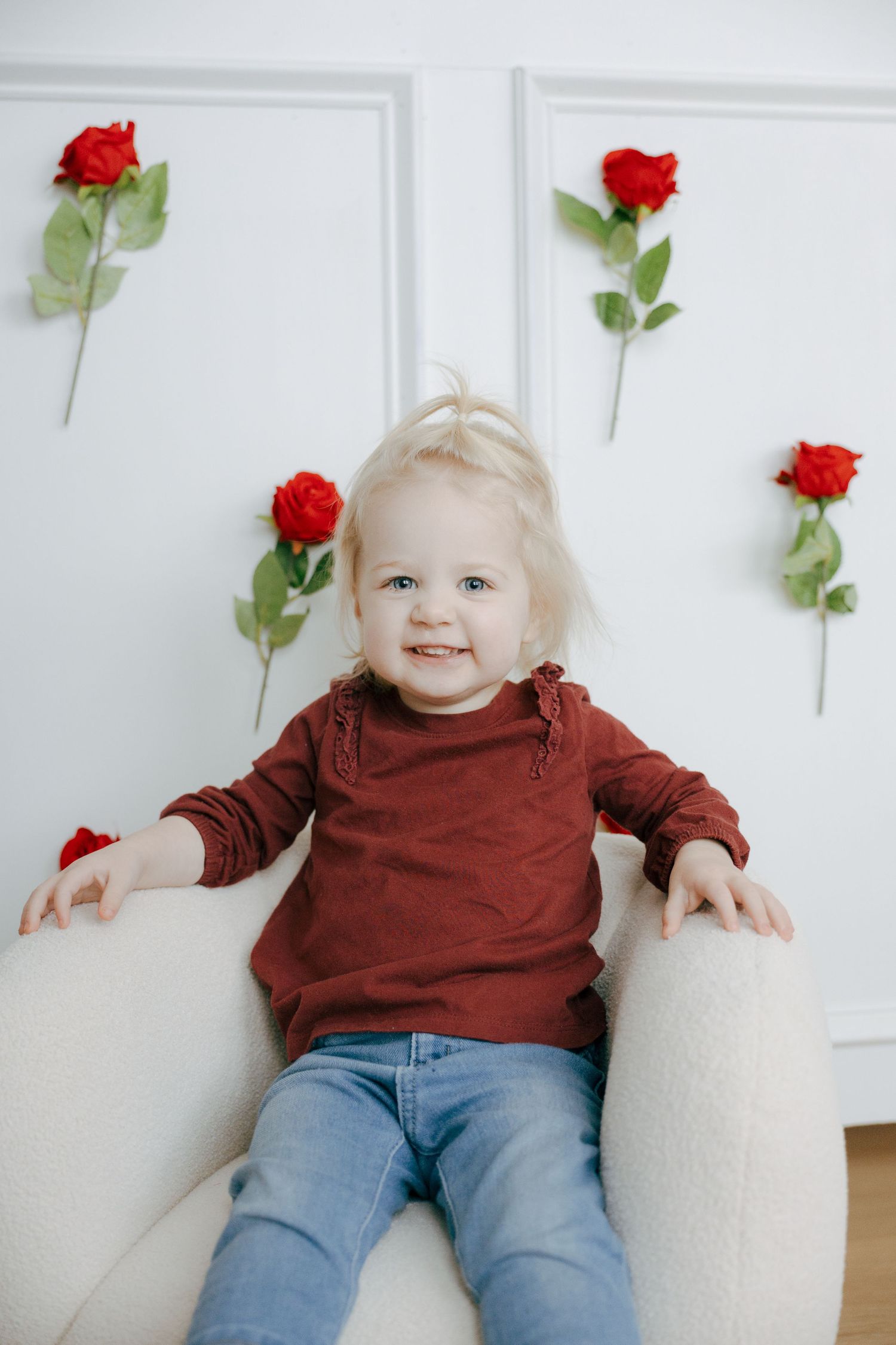 A small child in a burgundy sweater and jeans sits against a white wall decorated with red roses.