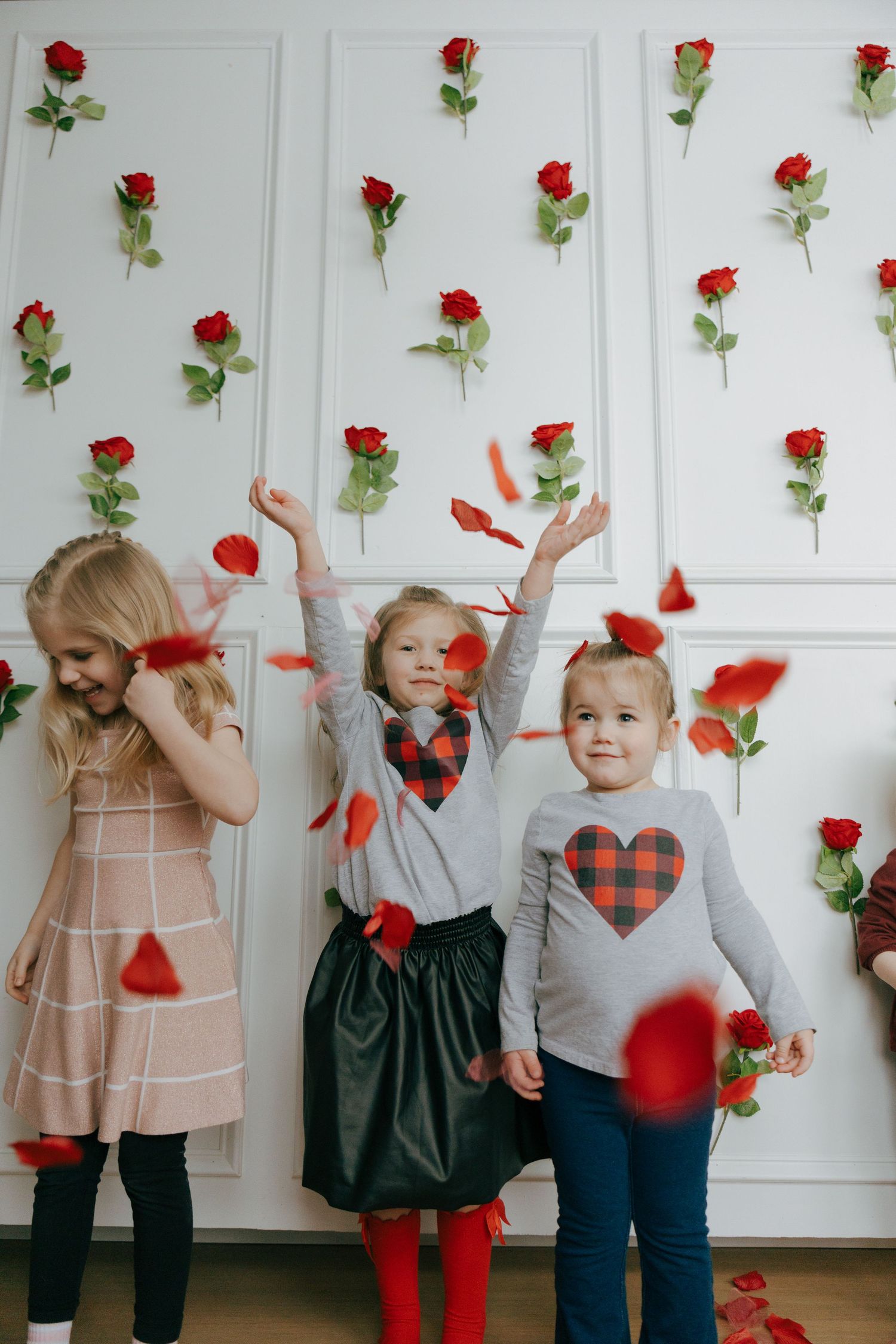 Red rose petals fall around children in winter outfits against a white wall decorated with single stem roses.
