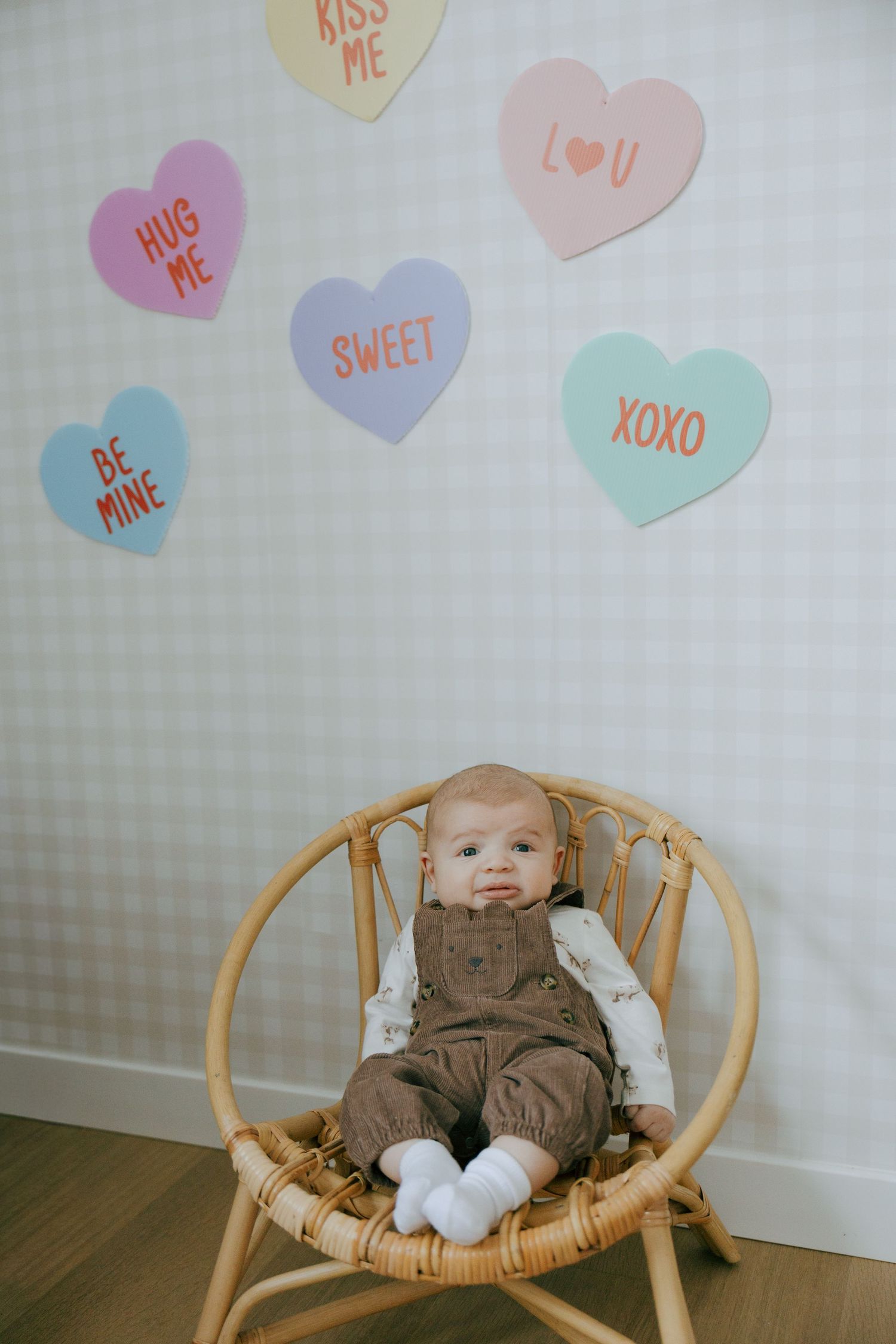 A baby sits in a rattan chair beneath colorful conversation heart decorations on a light gray wall.