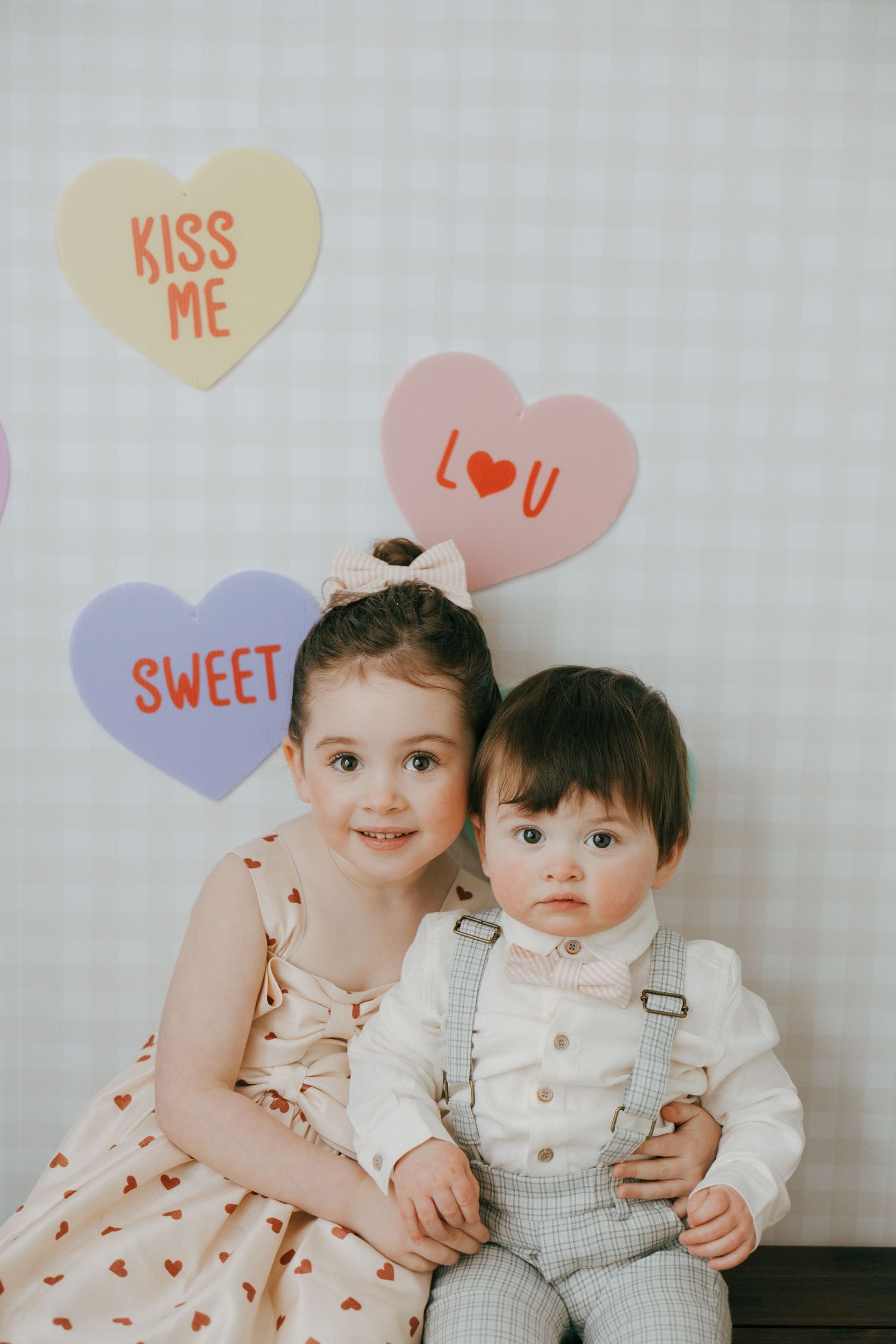 Two small children sit together against white wall with heart-shaped speech bubbles displaying sweet messages above them.