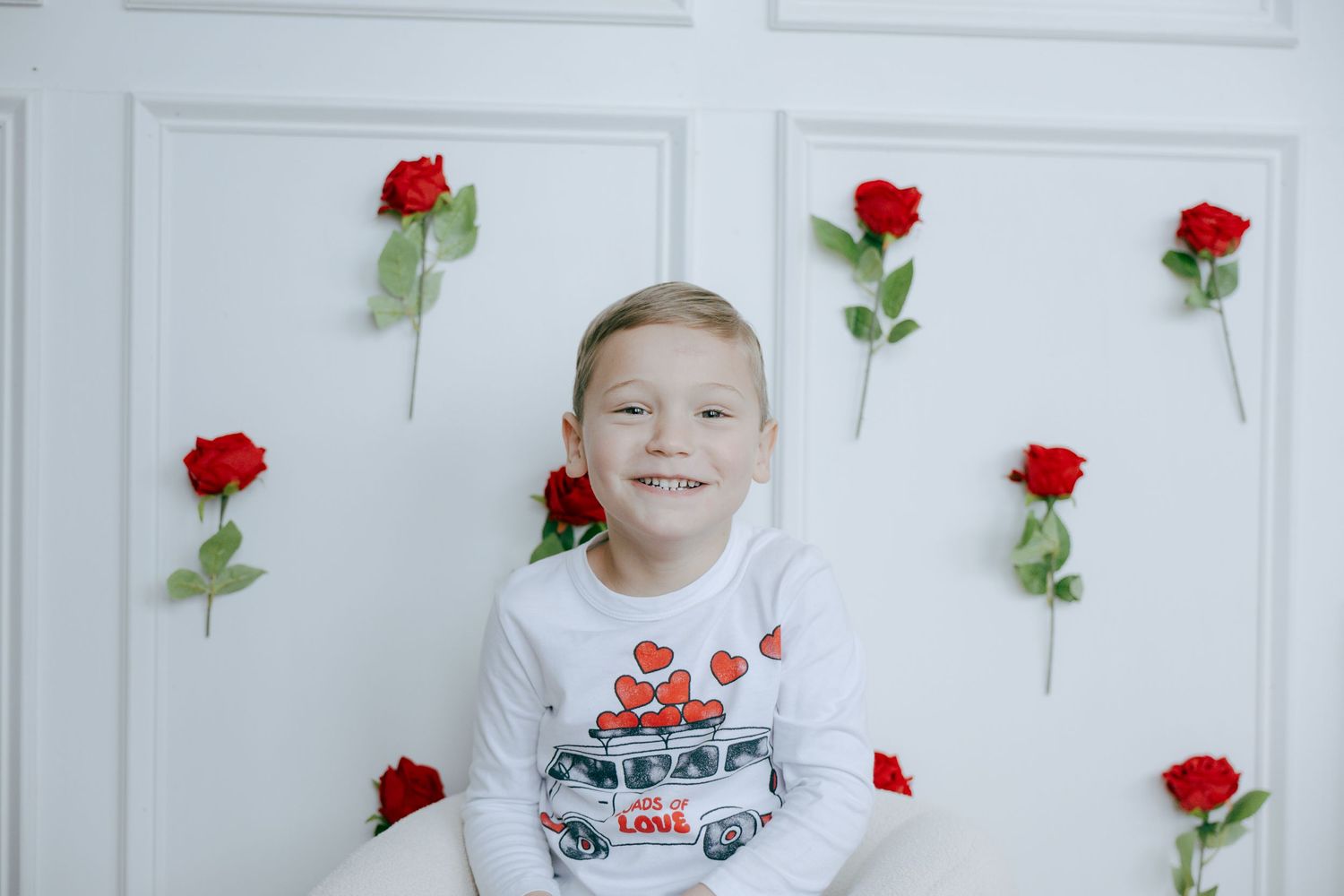 A white backdrop decorated with red roses features a child in a white and red outfit posing for a photo shoot.