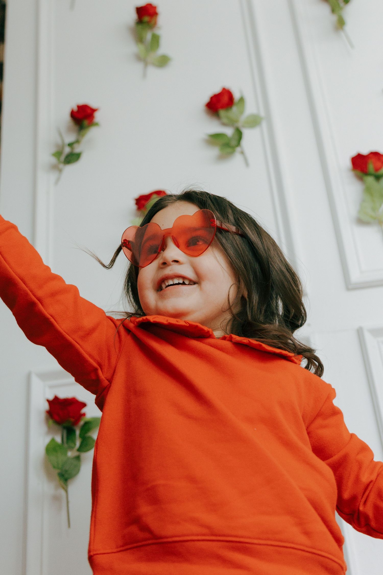 A person in a bright orange sweater dances joyfully against a white wall decorated with red roses.