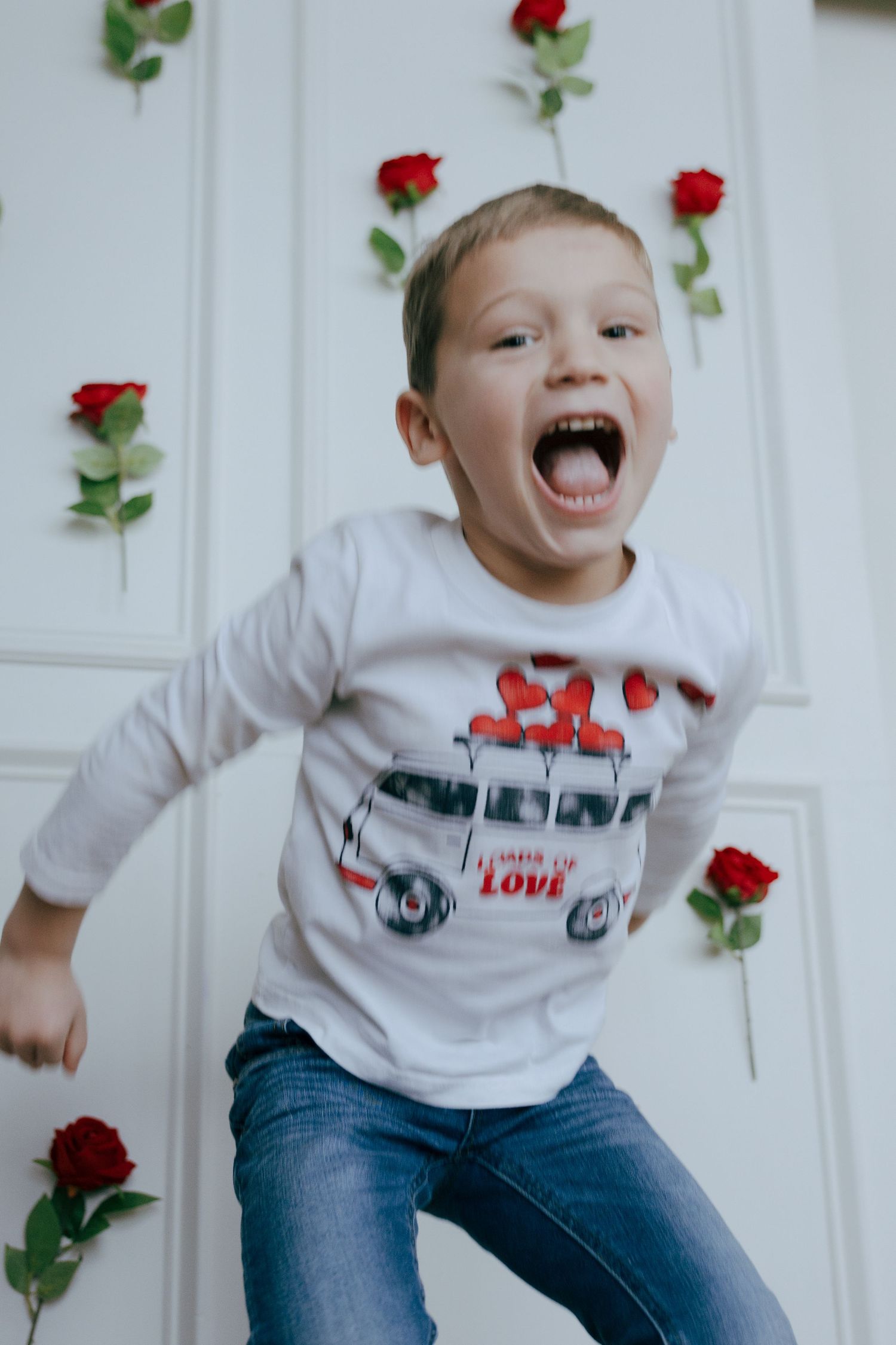 Energetic child wearing Valentine's themed shirt jumps joyfully against white wall decorated with red roses.