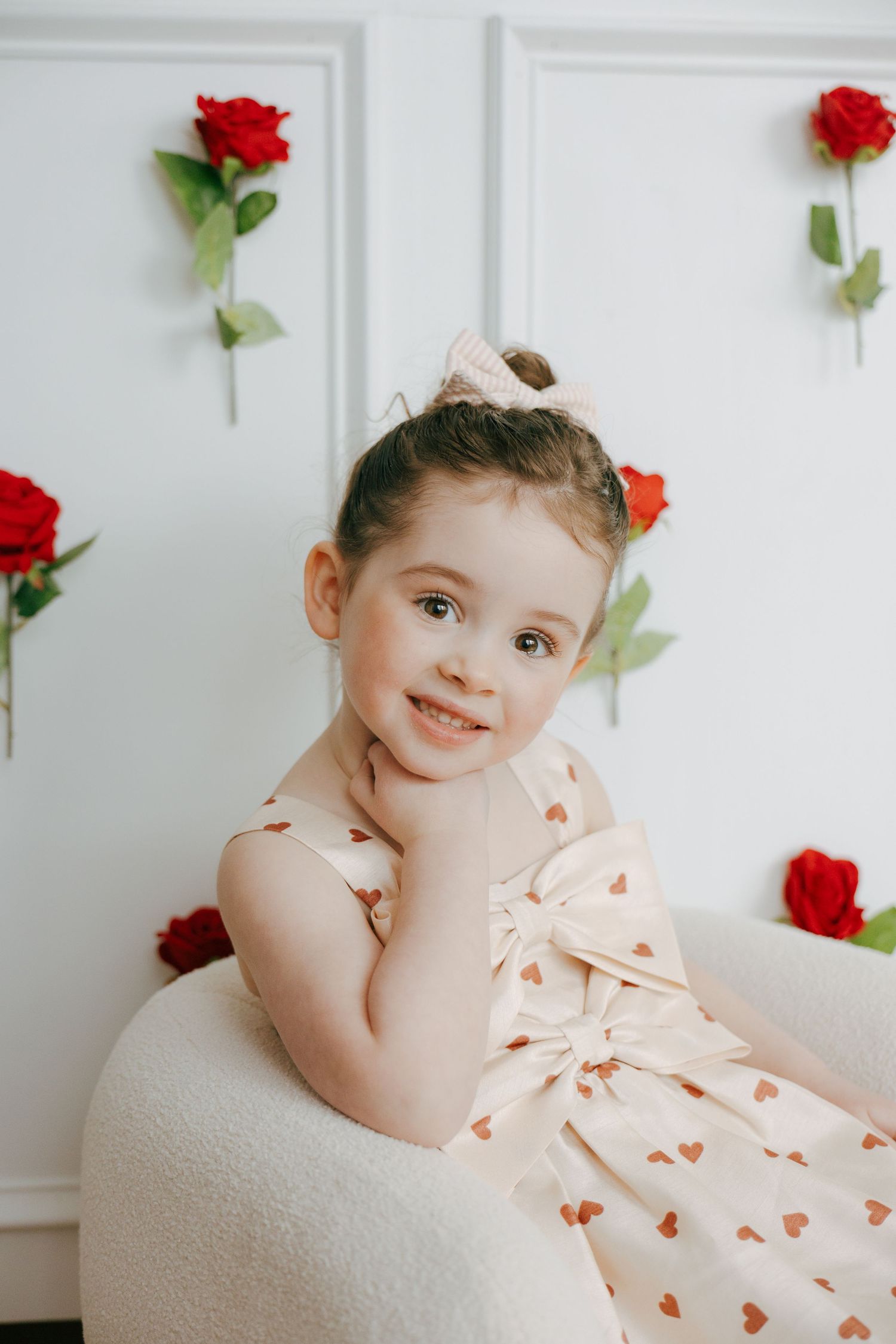 A child in a peach dress with heart patterns poses on a white backdrop with red roses.