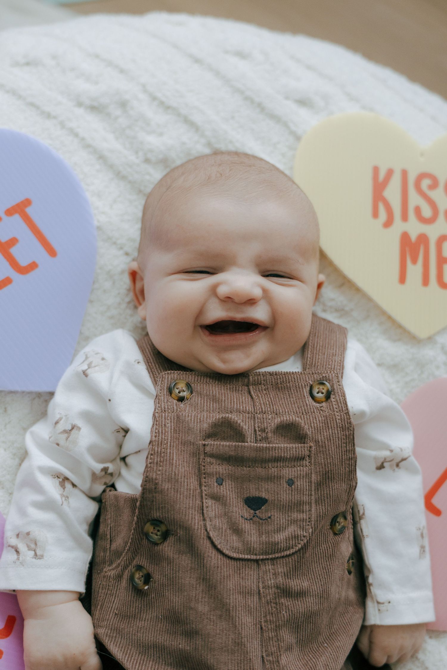 Smiling baby wearing brown overalls and white shirt lying on soft carpet with colorful decorations.