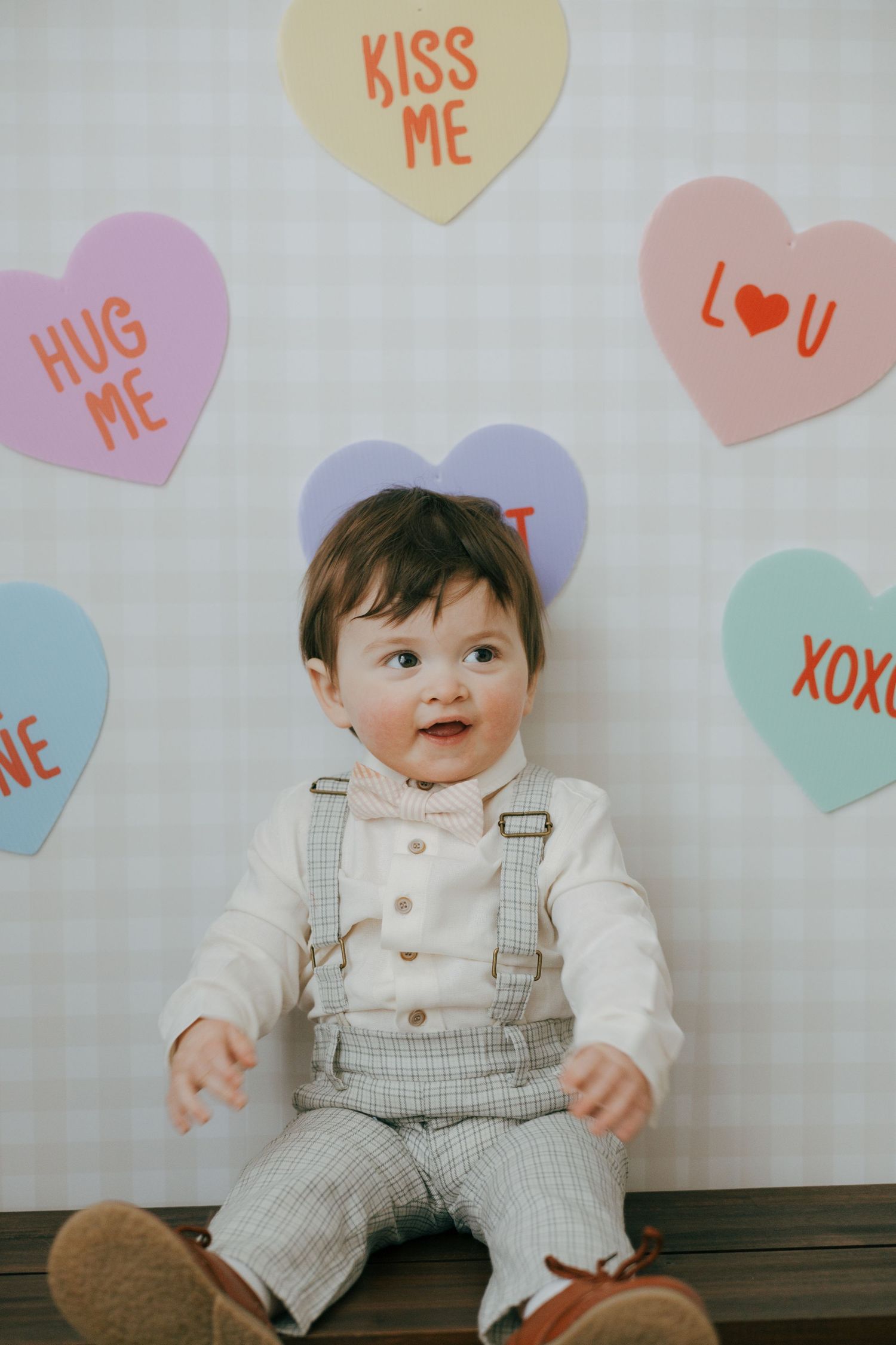 Baby wearing white shirt and plaid overalls sits against wall decorated with colorful heart-shaped Valentine's Day decorations.