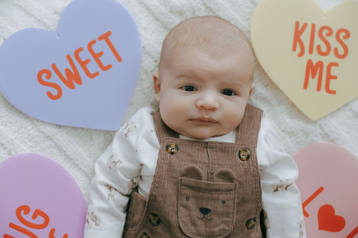 A baby wearing brown overalls sits among pastel colored Valentine's Day heart decorations and messages.