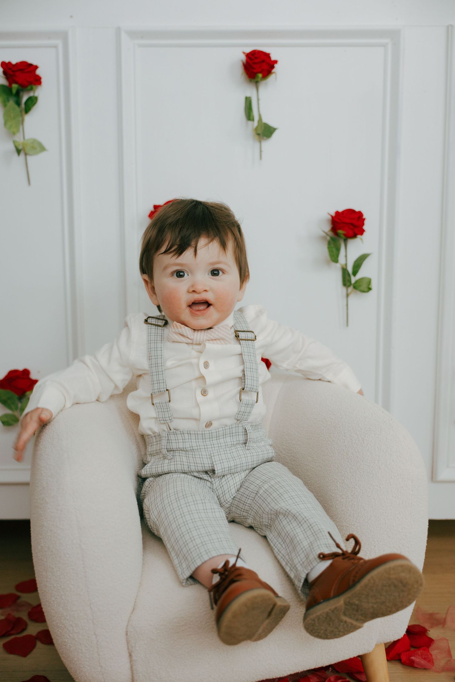 Small child in white overalls and brown shoes sits on cream-colored couch with red roses on white paneled wall.
