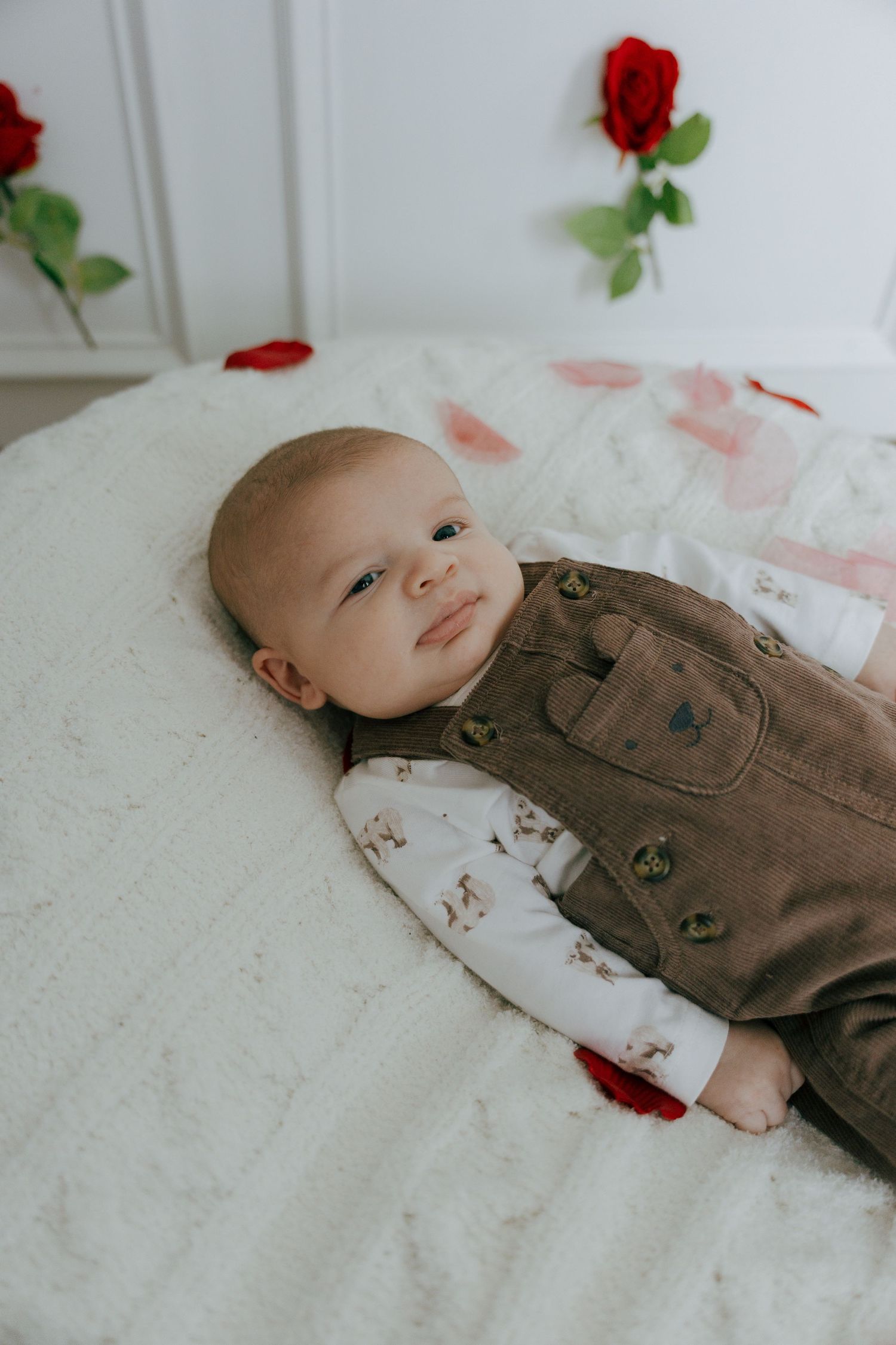 A baby lies on a white blanket wearing brown overalls with red rose petals scattered around.