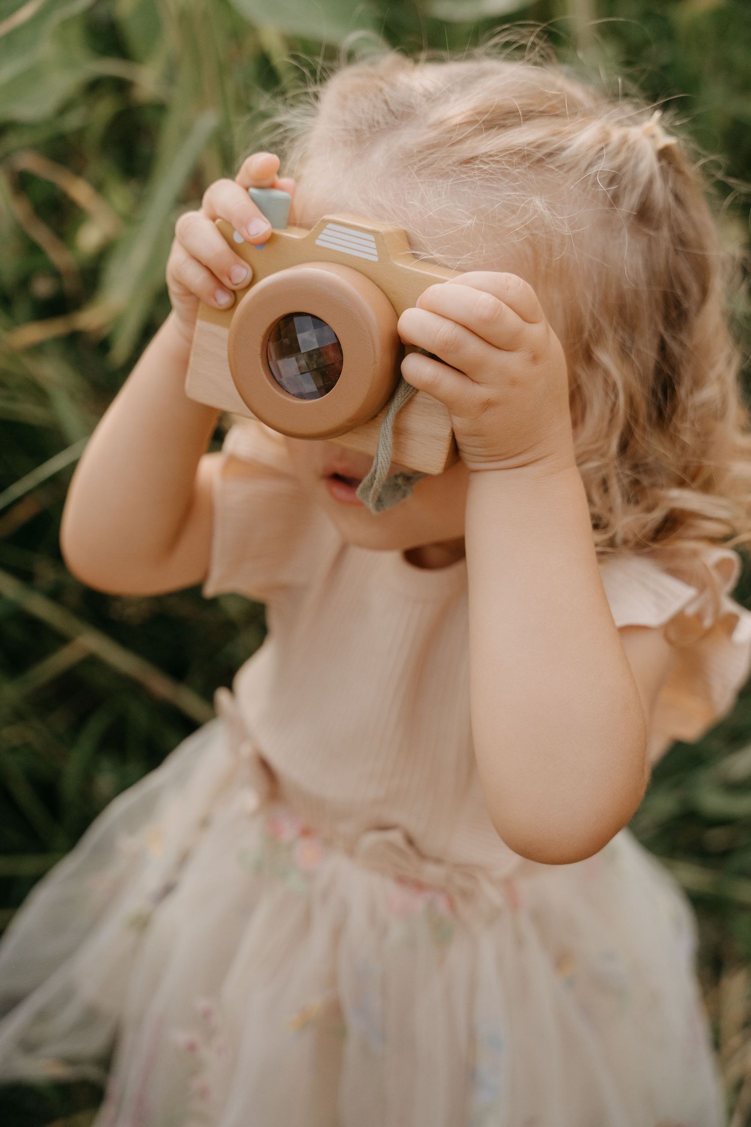 A child in a tulle dress holds up wooden toy binoculars to their eyes while playing outdoors.