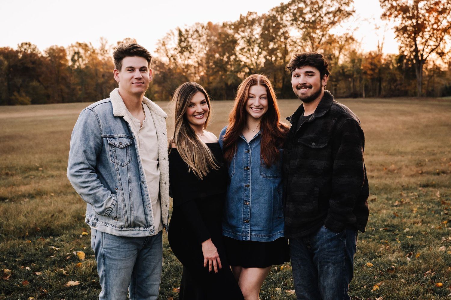 adult children pose together outdoors in casual denim and dark clothing during golden hour sunset in a grassy field. Family photographer in