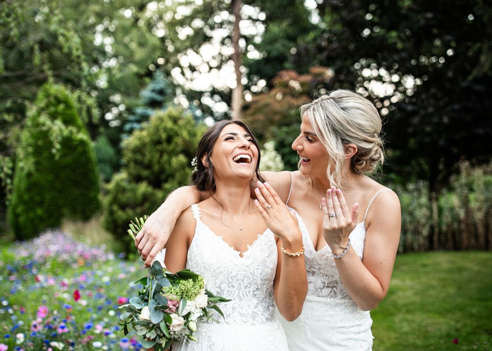 Two brides in white lace gowns laughing joyfully while showing wedding rings in lush garden setting.
