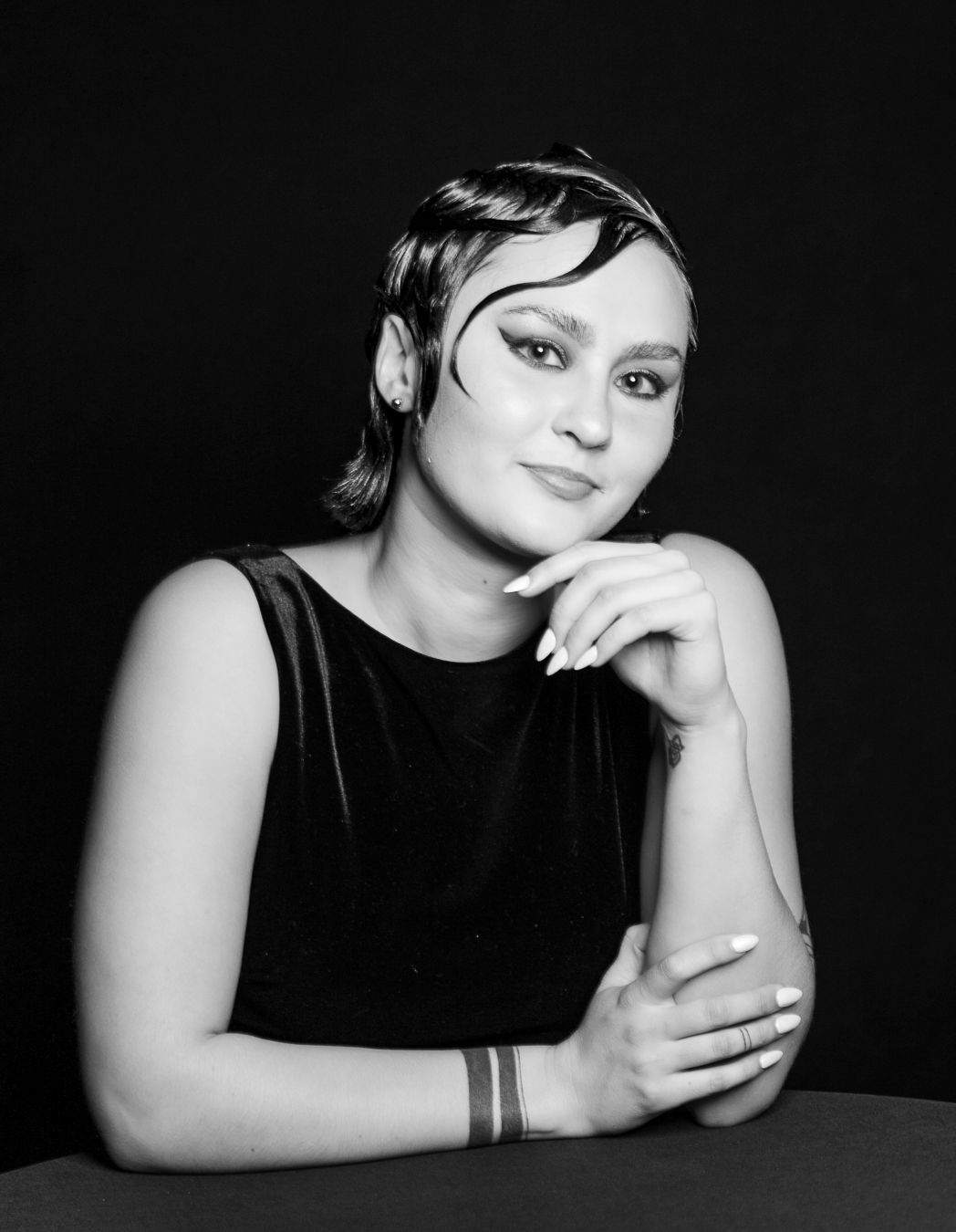 Black and white portrait of a person with short dark hair wearing a sleeveless black top against a dark background.