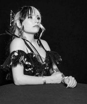 Black and white portrait of a woman in a dark ruffled dress seated at a table with a dramatic lighting effect.