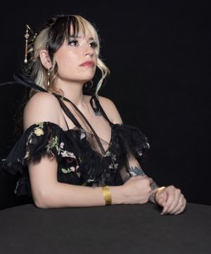 A person in a black floral dress sits thoughtfully at a dark table in a dimly lit setting.