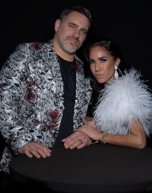 Couple posing together with one person in floral print jacket and other wearing white feathers.