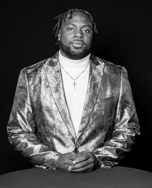 Person wearing shimmering metallic suit jacket seated against black background.