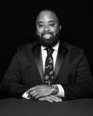 Professional black and white portrait of a smiling businessman in a suit and tie sitting at a table.