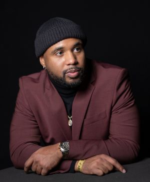 Person in burgundy suit jacket and beanie poses against dark background.