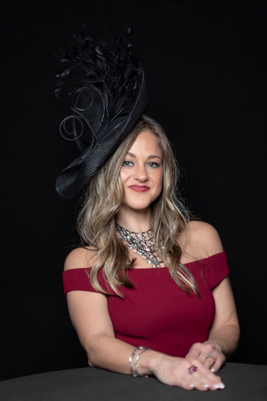 A woman in a burgundy off-shoulder dress and black fascinator poses elegantly against a dark background.