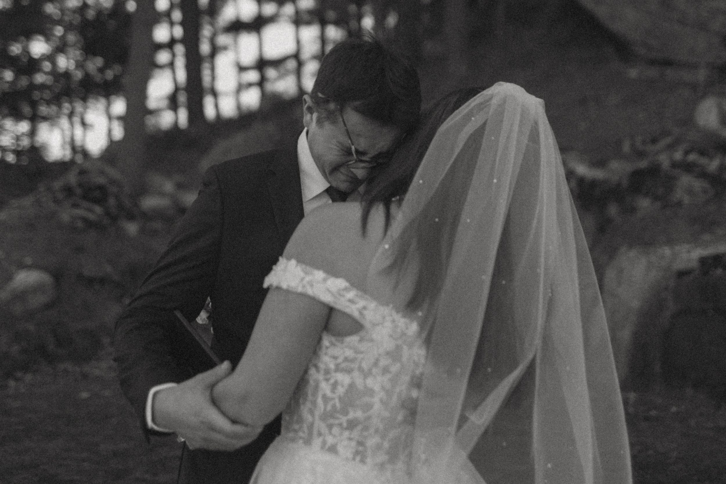 A wedding dress hangs in silhouette against a window in a moody black and white photograph.