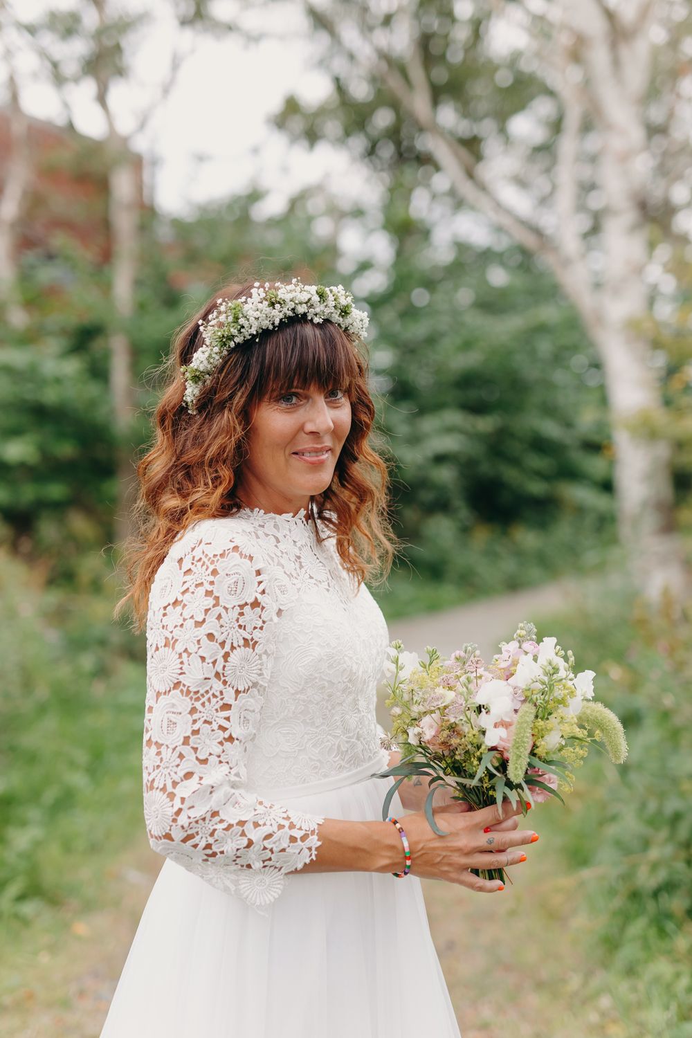 A bride in a lace-sleeved white wedding dress and floral crown holds a delicate bouquet in an outdoor garden setting.