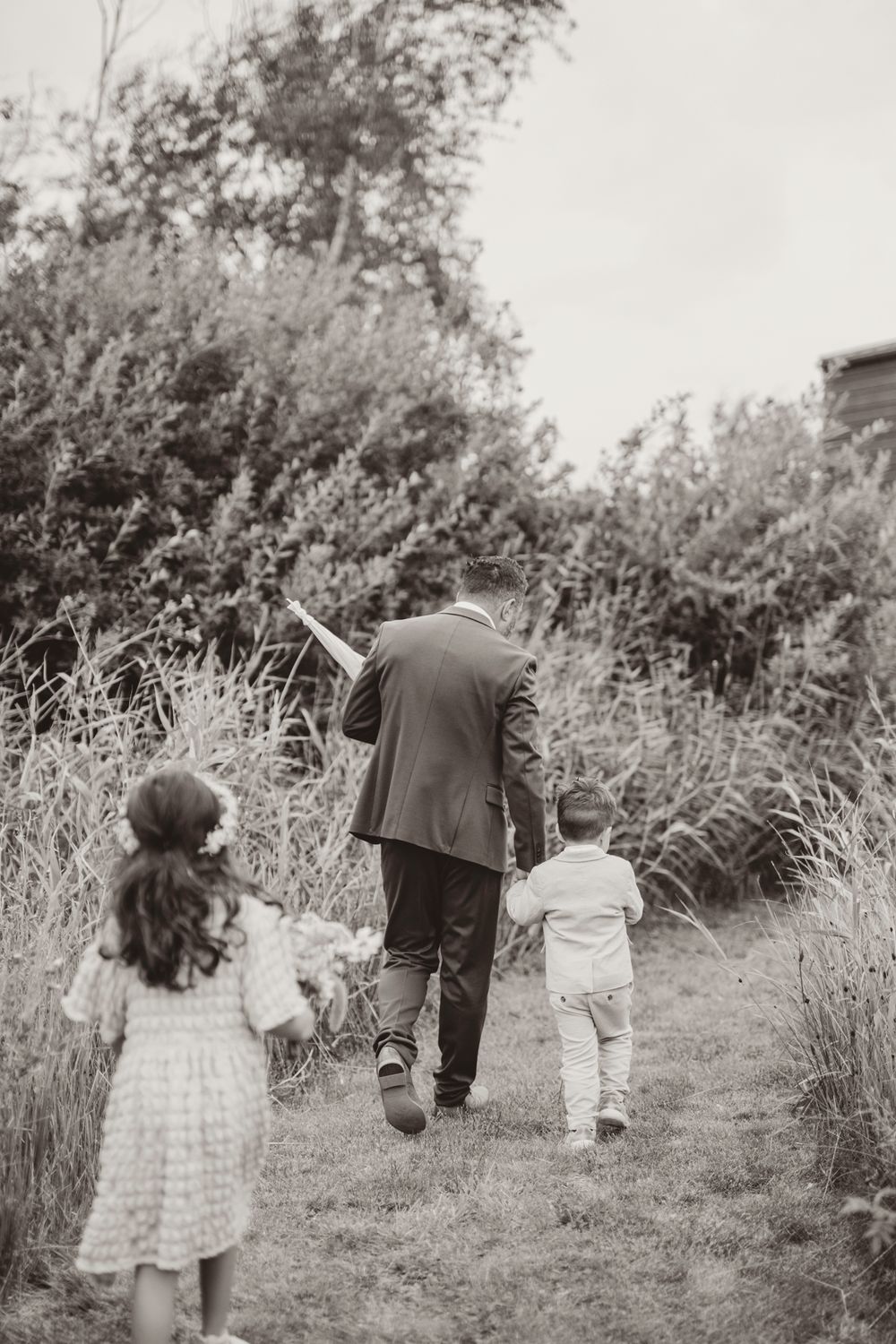A family walking away on a nature path together in black and white, holding hands through tall grass.