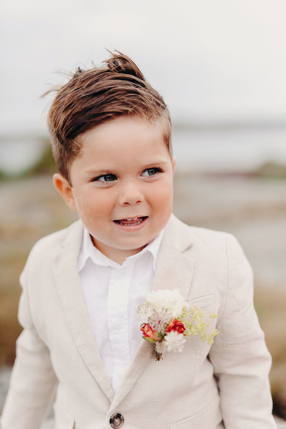 A young ring bearer in a beige suit and white bow tie smiles during an outdoor wedding ceremony.