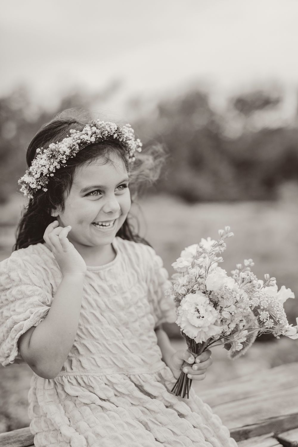 Black and white portrait of a child wearing a flower crown and textured dress while holding a floral bouquet outdoors.