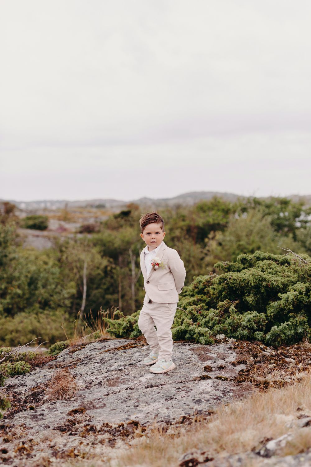 A child in a beige outfit stands on a rocky outcrop surrounded by green coastal vegetation on an overcast day.