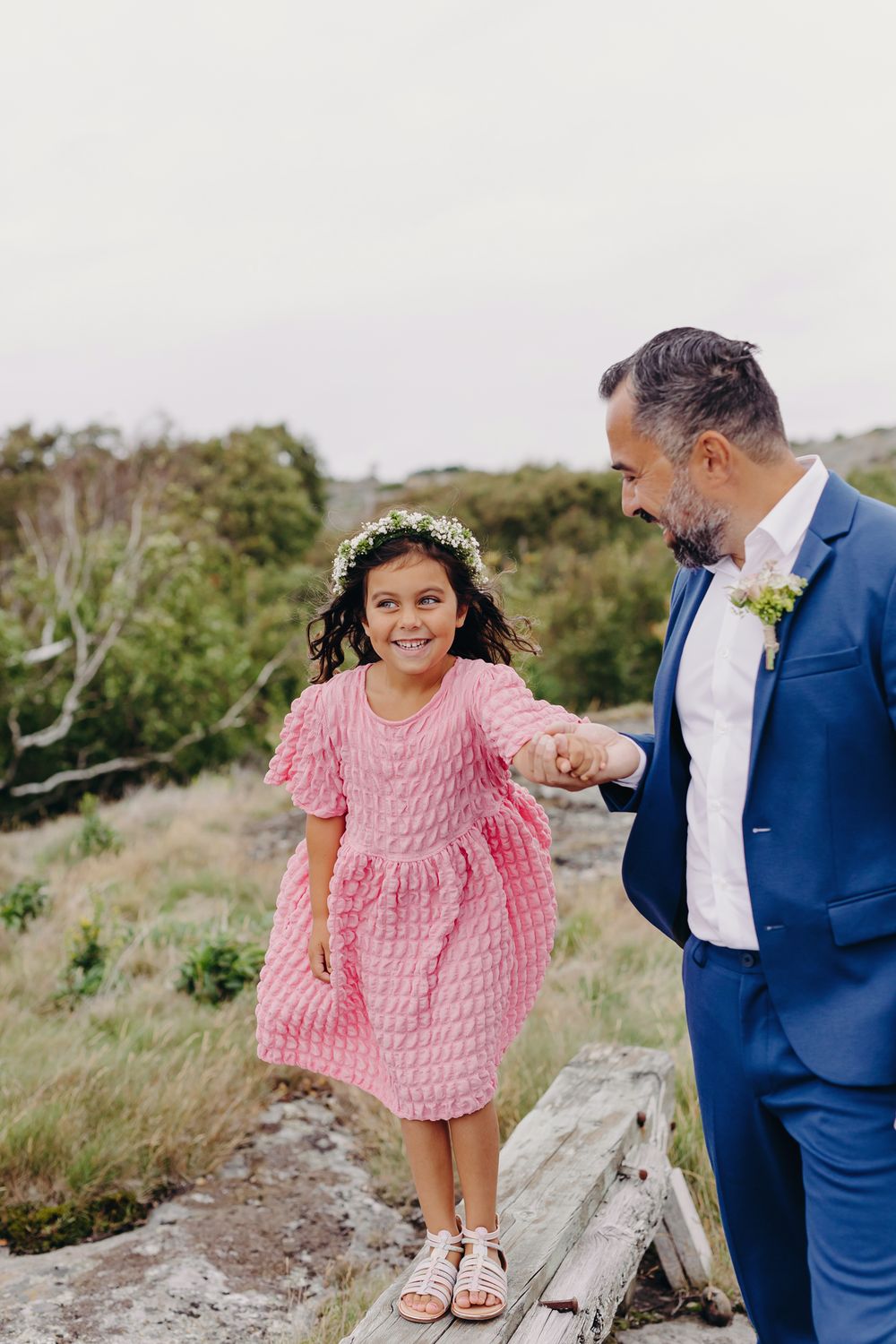 A child in a pink dress skips playfully along a rocky path while accompanied by someone in a blue suit.