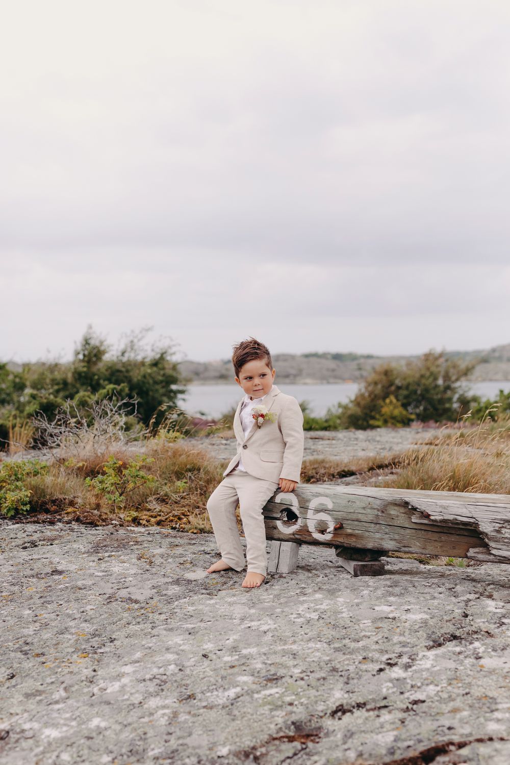 A child in beige clothing sits thoughtfully on a rocky coastal landscape overlooking the water on a cloudy day.
