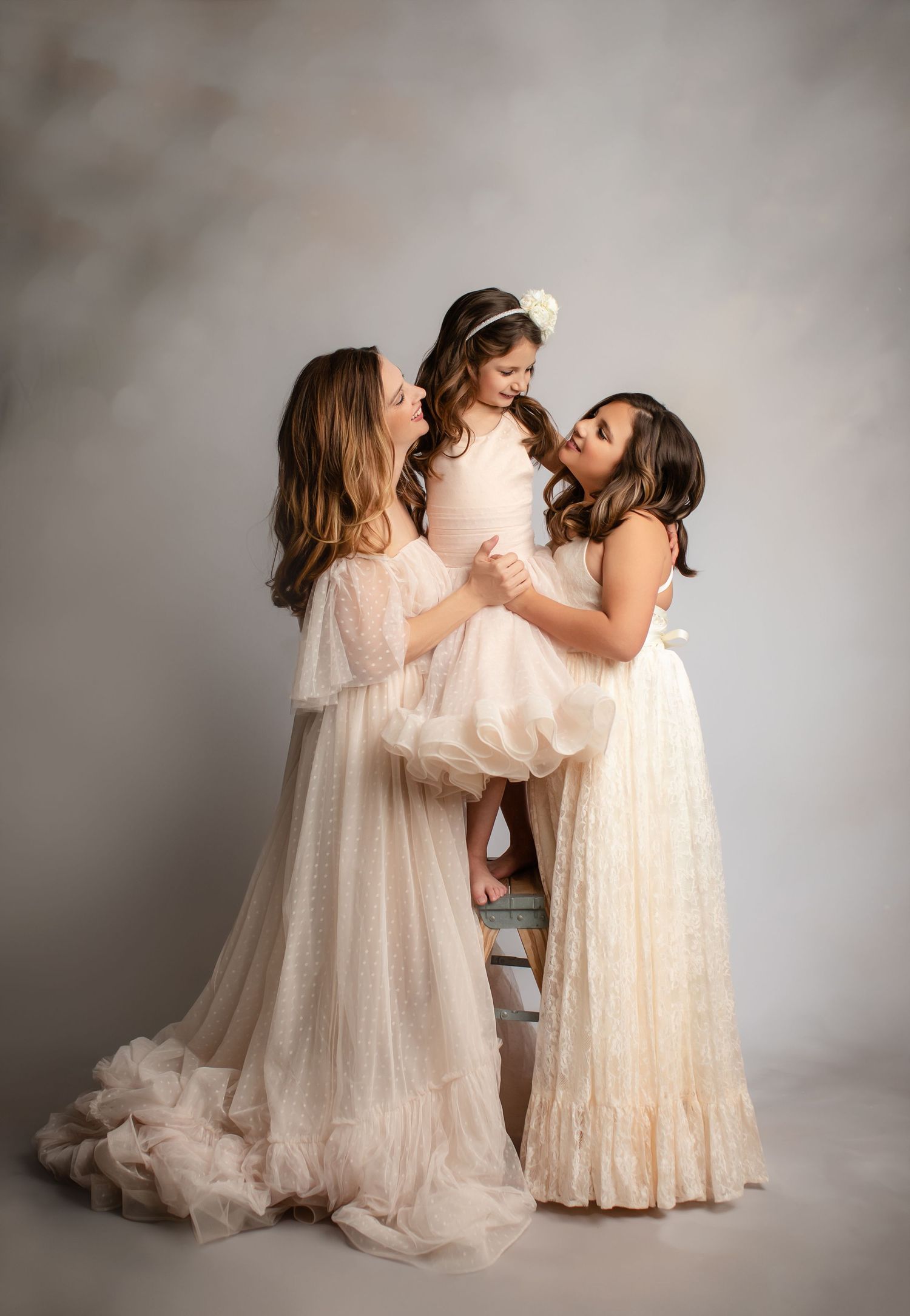 Three figures in flowing cream-colored gowns share an intimate moment against a gray studio backdrop.