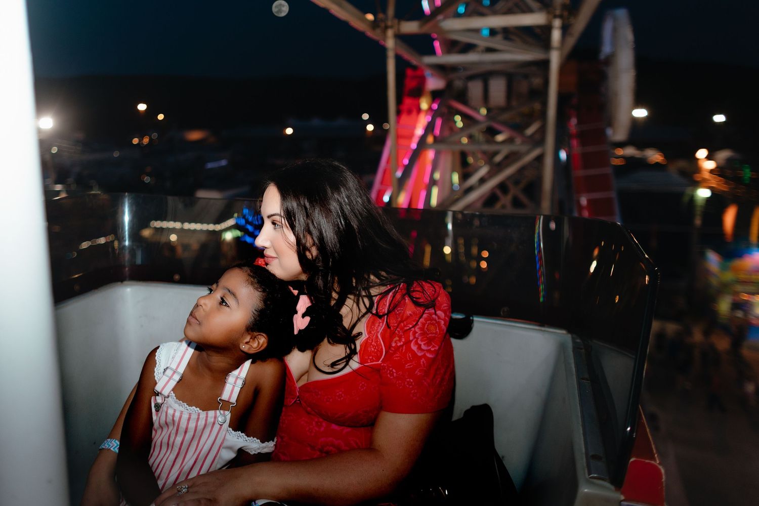Two people sit together on an amusement park ride at night, with colorful lights visible in the background.