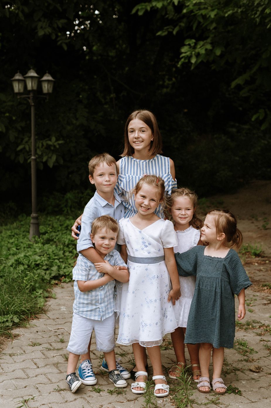 A group of young children standing together outdoors on a stone path surrounded by greenery.
