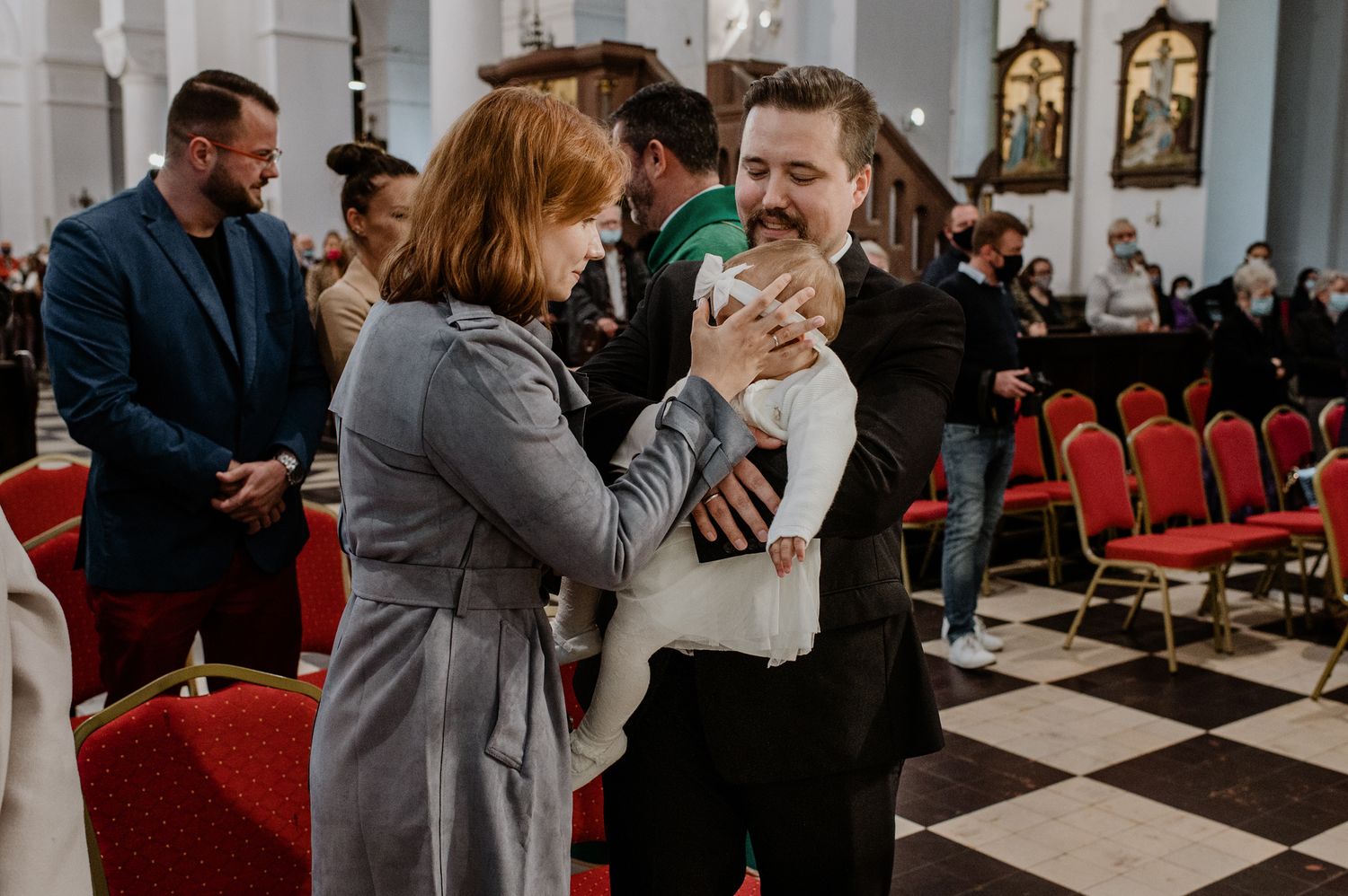 A couple dances together in a church hall with red chairs and checkered floor tiles.