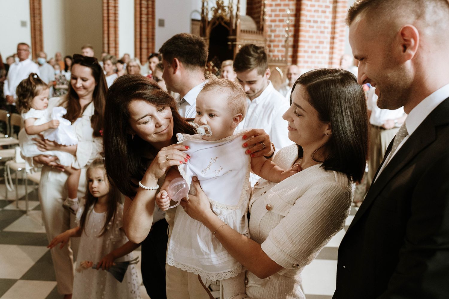 A joyful group gathers around a baby during a christening ceremony in a church.